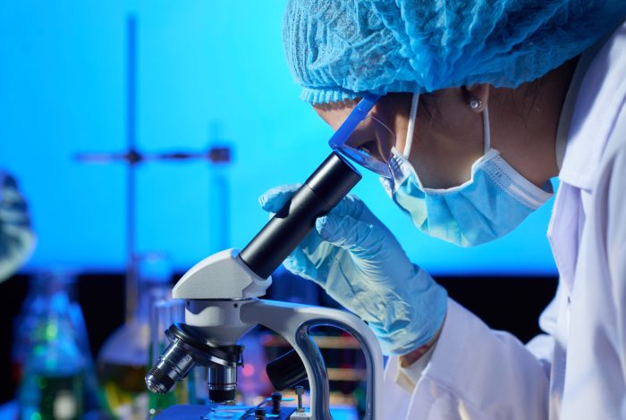 Examining Sample with Microscope Profile view of concentrated Asian microbiologist examining sample with help of modern microscope while wrapped up in work at dim laboratory