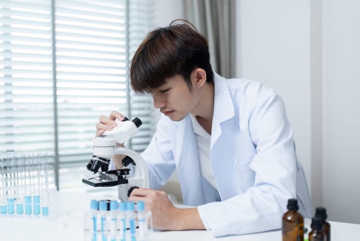 Young Researcher Using Microscope in Laboratory for Medical Drug Research and Development A young researcher in a lab coat uses a microscope in a modern laboratory, focusing on medical drug research and development.